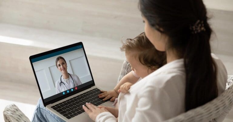 Mother holding infant child, using laptop to make video call to female pediatrician doctor